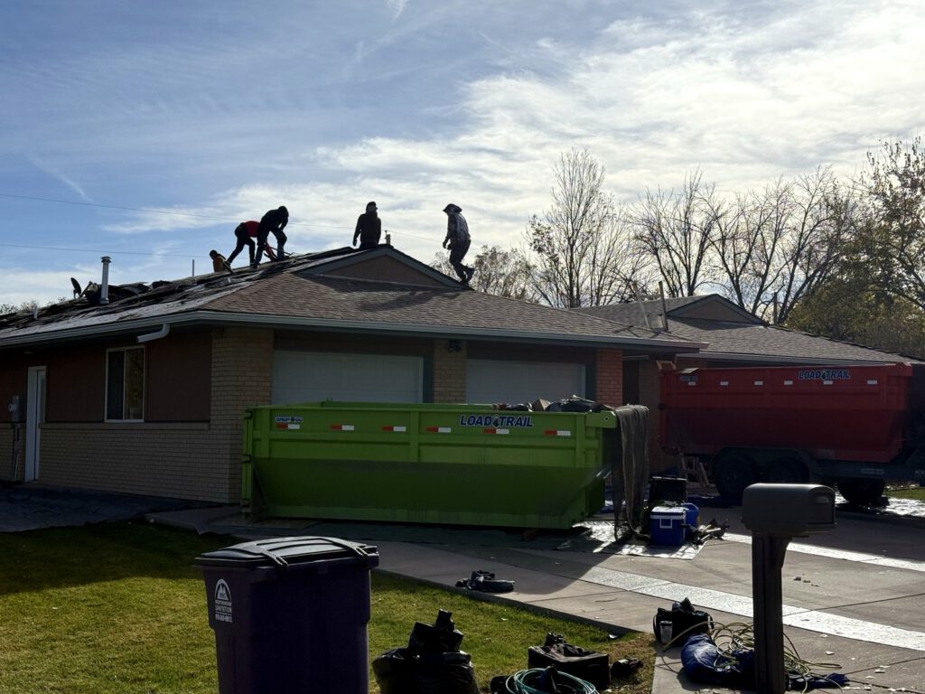 Roofing crew tearing off a storm-damaged roof, with a dumpster in the driveway