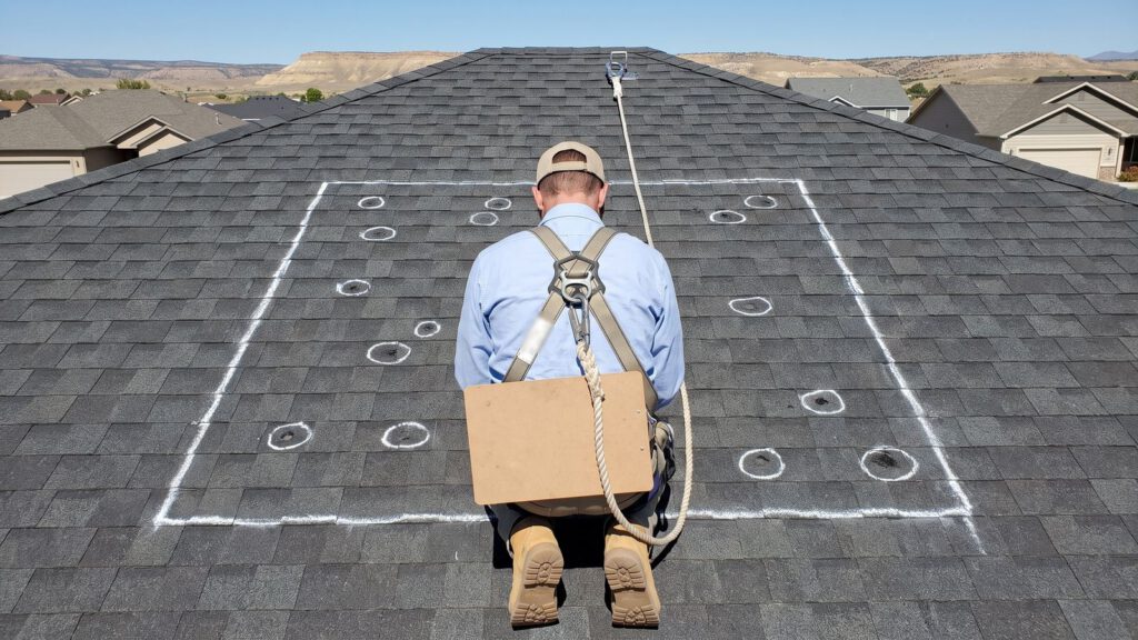 An insurance adjuster in a safety harness kneeling at a chalk-marked hail test square with small circled hail hits
