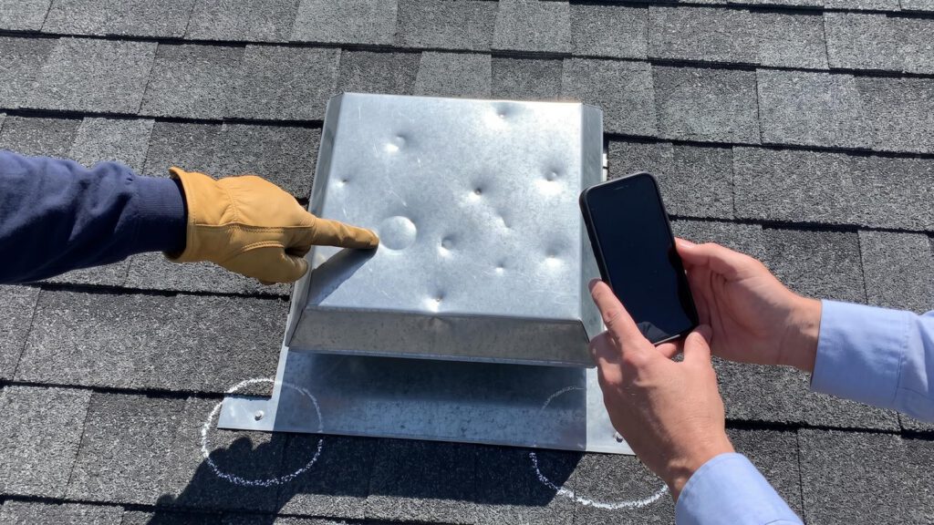 A contractor pointing at hail dents on a metal roof vent while the adjuster photographs them