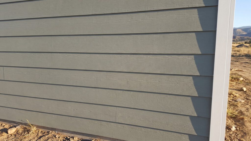 Close-up of new sage-gray fiber cement lap siding with an embossed wood-grain texture in low sun