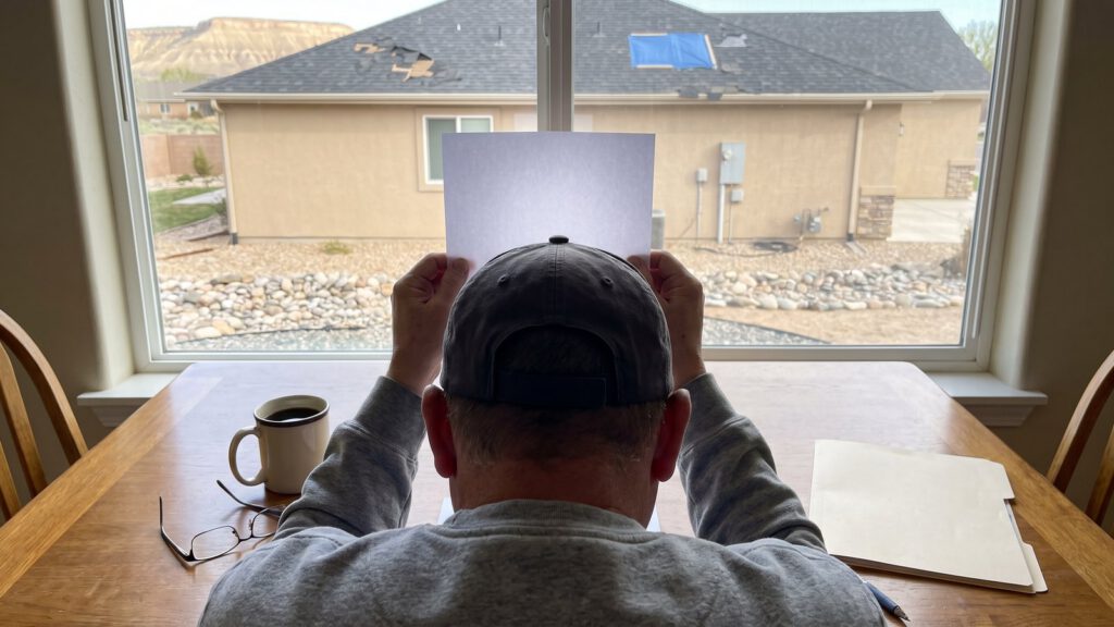 A homeowner at the kitchen table holding a paper up to the window, a hail-damaged neighbor roof outside