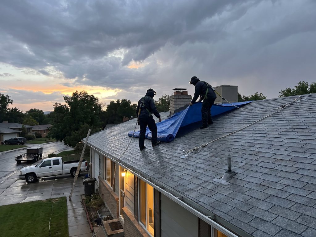 Roofers in harnesses spreading a blue tarp over a wet roof at dusk